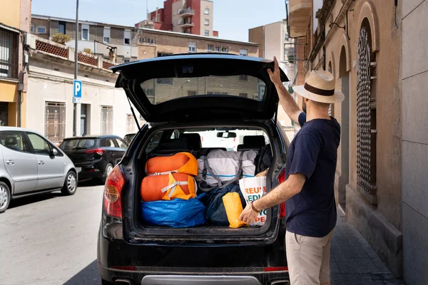 stock image unrecognisable man in a scout hat, T-shirt and shorts with his back turned closing a boot full of stuff for holiday travel and camping with sleeping bags, tent and clothes bag.