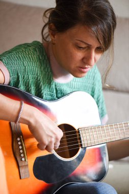 Kadın kanepede gitar öğreniyor, akustik enstrüman çalmaya odaklanıyor. Kırklı yaşlarda yeşil kazaklı bir kadın notalara bakıyor..