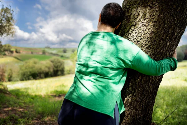 Non-binary woman with very short hair hugging a large oak tree in a field, green T-shirt and hair that is beginning to graying.
