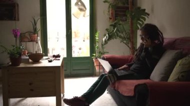 Rastalı, gözlüklü ve piercingli 40 'lı yaşlarda bir kadın, beyaz, telefonda telefonda, renkli örtülerle konuşuyor..