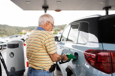 eighty-year-old man putting 95 petrol in a light blue car, short-sleeved green and white striped T-shirt, jeans and mobile phone case with mobile phone attached to the belt clipart