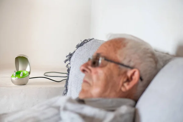 hearing aid for deafness in charger on white dining table, unfocussed older man in his eighties on sofa wearing glasses
