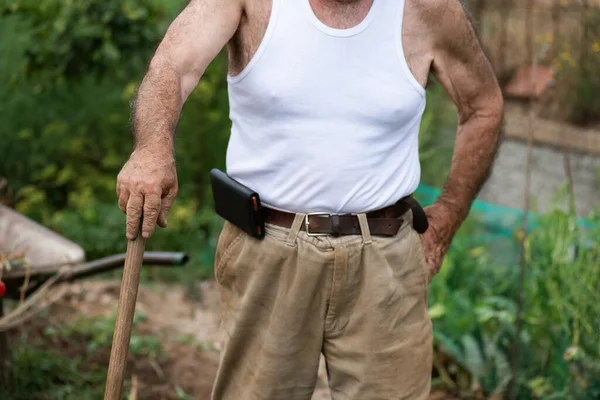 果园里一个认不出来的人 一个80多岁的退休男子 他的油箱顶部靠在锄头上 灯芯绒裤子 带着皮套的手机 — 图库照片