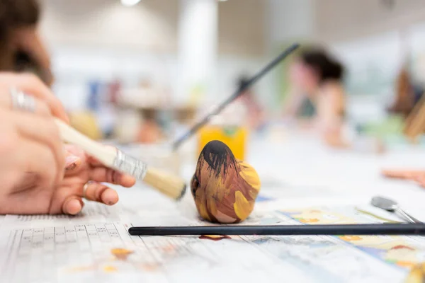 stock image detail of a woman's hand with a brush painting a ceramic piece in a workshop with other people learning, painting of oxides and minerals on a clay bird.