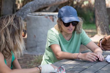 Yeşil tişörtlü, güneş gözlüklü, tatil pikniğinde sohbet eden ve hayatı ve açık hava maceralarını anlatan iki arkadaş..