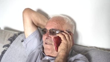 80 'li yaşlarda bir adam cep telefonuyla konuşuyor. Kanepede, gri saçlı, güneş gözlüklü, kahverengi tenli. Çizgili gömlek