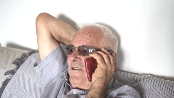 80多岁的男人在沙发上用手机聊天 灰白的头发 太阳镜 屋里的棕色皮肤 条纹衬衫 — 图库视频影像