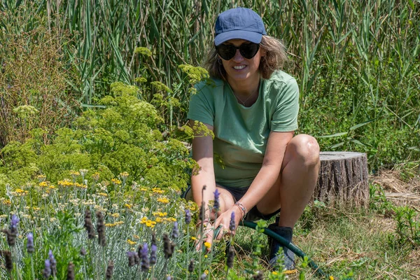 浇灌蔬菜园的女人蹲伏下来 看着镜头笑着 穿着短裤 绿色T恤和帽子 花园里种着芬芳的植物 — 图库照片