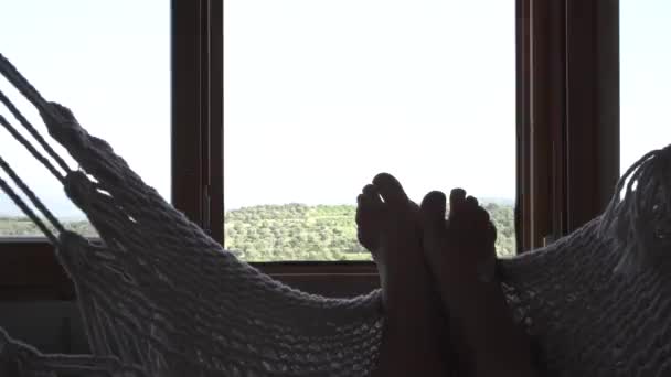山や森や畑の景色を望む大きな窓の中で マクラムのスイングでリラックスした女性の足のビデオ — ストック動画
