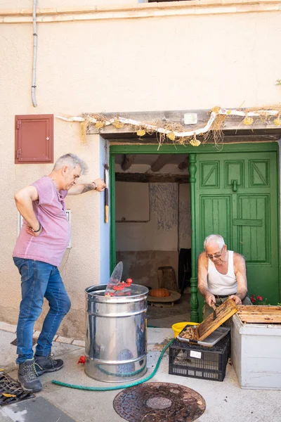 older man cutting the wax from the bee panels at the door of a house to extract the honey with the centrifuge, older man teaching his fifty year old son the process and beekeeping.