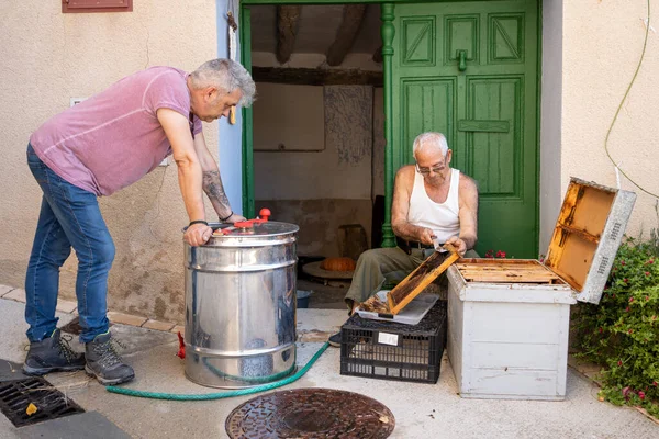 遠心分離機で蜂蜜を抽出するために家のドアの蜂のパネルからワックスを切断した老人50歳の息子にプロセスと養蜂を教える — ストック写真