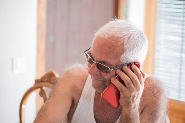 80 'li yaşlarda beyaz atletli yaşlı bir adam kestane rengi bir cep telefonuyla konuşuyor gri saçlı ve kahverengi tenli, gülümsüyor..