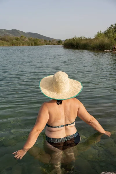 woman on her back bathing in river with bikini, chubby white-skinned person in her forties with straw hat with wide eaves, natural landscaping