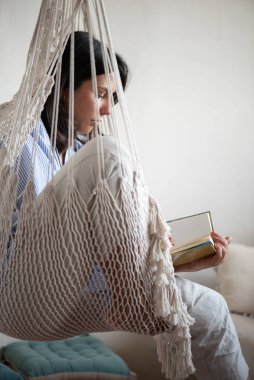 20 'li yaşlarında bir kadın lüks bir dairede makrome bir hamakta kitap okuyor, rahat bir kız krem rengi edebiyat okuyor ve eğleniyor.