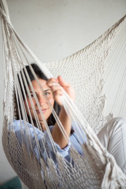 Bir kadın makrome bir hamakta oturmuş tasarımcı iç çamaşırlarıyla, günlük mavi çizgili bir gömlek giymiş, makrom ipliklerden bakıp gülümsüyor..