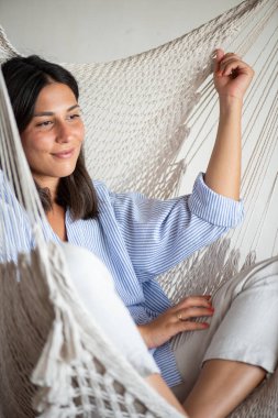20 yaşında bir kadın, makrome bir hamakta, tasarımcı iç tasarımında, günlük mavi çizgili bir gömlek giymiş, sonsuzluğa bakıyor, mutlu, düşünüyor.