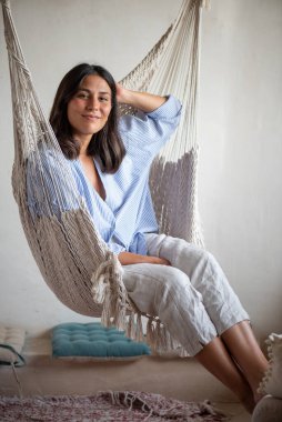 20 yaşında bir kadın, makrome bir hamakta iç tasarımcı olarak oturuyor, günlük mavi ve beyaz çizgili bir gömlek giyiyor. Doğal ışık ve konforlu alan