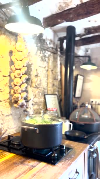 火に沸騰するポット 冬の雰囲気のキッチン 石の壁が付いている田舎スペース 壁に掛かるニンニクの大きいひも 野菜を沸かした大きな鍋 金属スポットライト — ストック動画