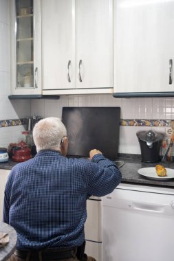 Mutfaktaki tabureye oturmuş, sırtı dönük, tanınmaz halde yaşlı bir adam. Mavi kareli gömlek, panama pantolon, gri saç ve kulağında işitme cihazı. Meyve sınırlı beyaz mutfak.