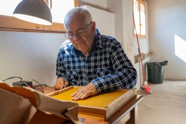 Yaşlı arı yetiştiricisi çerçeveye küçük trafolar ve elektrik temasıyla balmumu yapıştırıyor. Üzerinde mavi-beyaz bir tişört vardı. Atölyede bir pencerenin yanında, lamba yanıyordu.