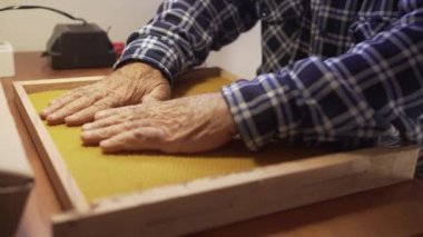 Eski bir arı yetiştiricisinin yıpranmış elleriyle küçük bir trafoyla çerçeveye balmumu çarşafları yapıştırmasının ayrıntıları..