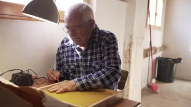 Yaşlı arı yetiştiricisi çerçeveye küçük trafolar ve elektrik temasıyla balmumu yapıştırıyor. Üzerinde mavi-beyaz bir tişört, atölyede bir pencerenin yanında, işi aydınlatan bir lambayla..