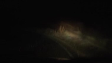 Araba virajları olan gizemli ve misafirperver olmayan bir yolun videosu, arabanın farları tarafından aydınlatılan, ön camları açık yağmurlu bir gece.