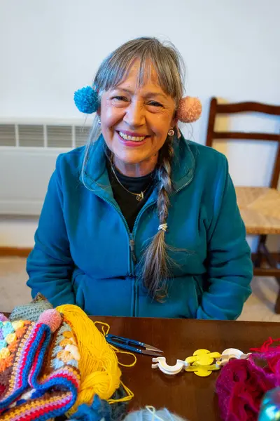 Woman with pompoms over her ears laughing. Person in her sixties with long grey hair and a long plait. Dressed in a blue fleece. Interior space with chair and electric hotplate, woolens on table.