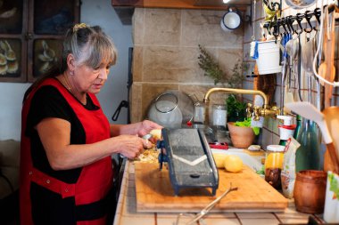Kırsal mutfakta yemek pişiren bir kadın, 60 'larında emekli, uzun gri saçlı, kırmızı önlüklü, ahşap üzerinde patates kesen bir kadın..