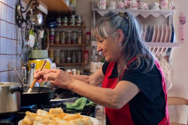 Kırsal mutfakta yemek pişiren, 60 'larında emekli, uzun gri saçlı, kırmızı önlüklü, gülümseyen ve kızartma tavasında ahşap kaşıkla karıştıran bir kadın..
