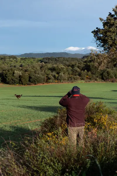 Sırtı dönük tanınmayan bir adam, köpek dürbünüyle tarlada koşan, dağ kıyafetleri giymiş, kahverengi pantolon giymiş, kafasında kestane rengi post ve fular olan köpeğe bakıyor..