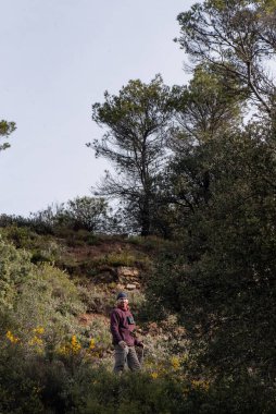 Ormanda dağ kıyafetleri giymiş bir adam, İskandinav sopaları, kestane rengi bir post ve kafasında yün bir ceket, yüzünde bir gülümsemeyle kameraya bakıyor.