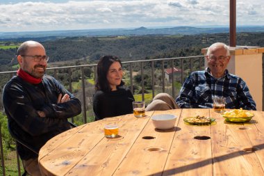 Üç nesildir ailesi terasta aperatif yiyor, torunu yirmili yaşlarda, babası kırklarında gülümsüyor ve dedesi kameraya bakıyor. Arka planda tarla ve orman manzaralı manzara.