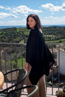 20 'li yaşlardaki kadın yüzünü kameraya çevirdi, tahta masa, yeşil sandalyeler, uzun siyah şallar, koyu renk saçlar, doğa manzarası ve manzara.