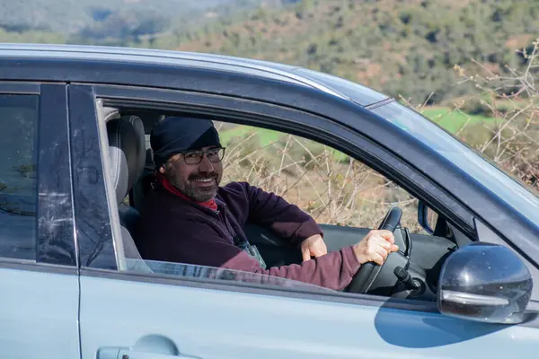 man at the wheel of a 4x4 car looking at the camera and smiling, image from outside the car. Light blue off-road car, we see the window and the driver with glasses, beard, wearing a polar jumper and a polar scarf on his head.