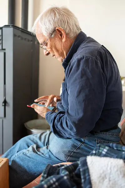 八十岁的男子坐在沙发上玩手机和冲浪 穿着牛仔裤和海军蓝灯芯绒衬衫 免版税图库照片