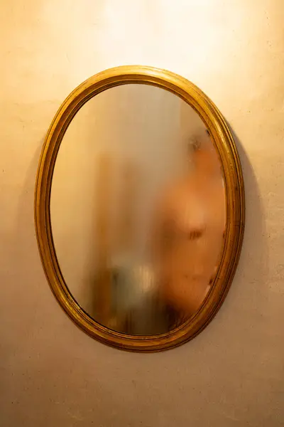 一个裸体女人和一个木制的结构与毛巾在一个雾蒙蒙的镜子的倒影与一个金色的框架 乳白色的墙精美的水泥 来自镜子上方的暖光 图库图片