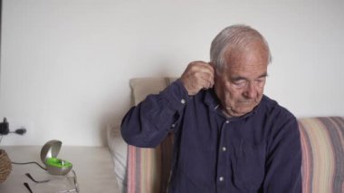 Yaşlı bir adam kanepeye oturur ve iki işitme cihazını takar, sonra da gözlüklerini takar. Lacivert gömlek
