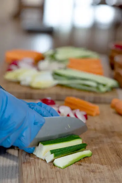 女人在木头上切萝卜 坐在厨房桌旁 绿色T恤 牛仔围裙 蓝色乙烯手套 图库图片