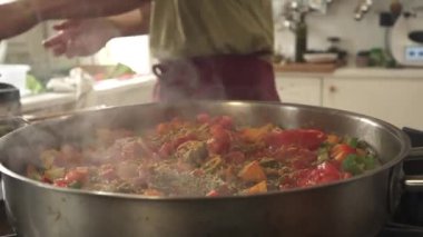 Bir adamın elinin büyük tavaya baharat koyduğu video detayı, endüstriyel kızartma tavası arkasında kestane rengi etek ve yeşil tişört, doğal ışık.