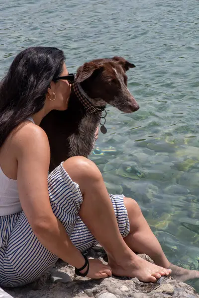 Güneş gözlüklü, uzun kahverengi saçlı, nehirde bir köpeği öpen, çizgili pantolonlu ve beyaz kolsuz bluzlu bir kadın..