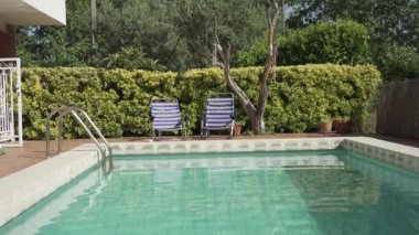 Yüzme havuzunun önünde boş mavi ve beyaz çizgili alçak hamaklar ve insansız mavi su, suyun hareketini görebildiğiniz ve size yazı hatırlattığı dikey video..