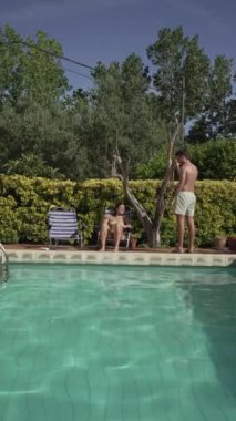Havuzun kenarında oynayan bir çiftin suya atlamak için oynadığı dikey video. Aşıkların yaz oyunları turkuaz suda serinliyor..
