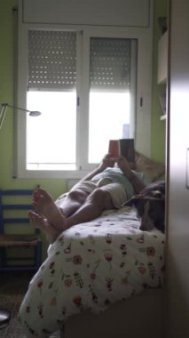 Yatakta köpeğinin yanında kitap okuyan bir adamın dikey videosu, ön planda çıplak ayaklar ve karanlık köpeğin yüzü sıkılmış ve gevşemiş. Fıstık rengi yorganlı gençlik odası.