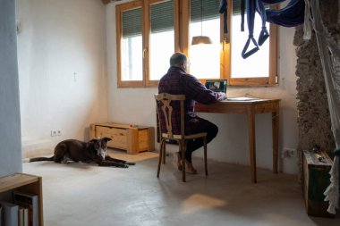 Kırsal bir alanda köpekli, pencerenin önünde, bilgisayarlı ve ipad 'li kırmızı ve mavi kareli gömlekli ve boynunda atkısı olan kel ve gözlüklü bir adamla tele-posta atıyor..