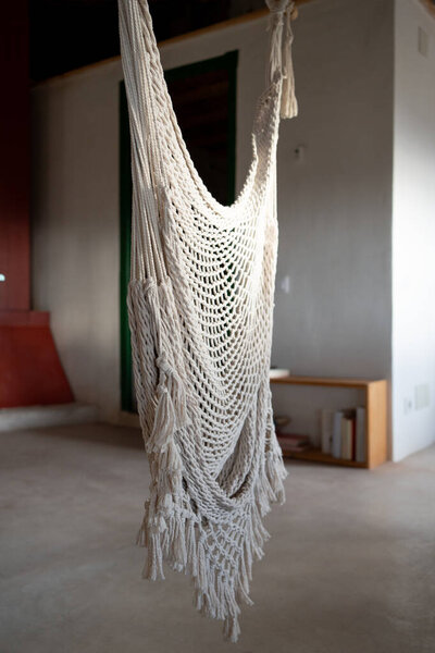 empty macrame hammock in the interior of a rustic house, bookshelf with books and iron chimney structure in the back.