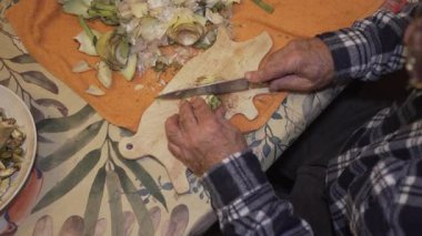 Yaşlı bir adamın, domuz şeklinde ahşap bir tahtanın üzerinde alkarchofa doğrayan zenital görüntüsü. Vejetaryen yemeğinde renkli bir sebze..