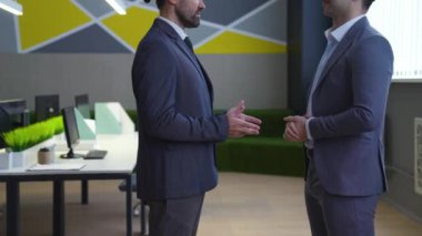 Ofiste takım elbiseli iki adam konuşuyor, gülümsüyor, el sıkışıyor, pazarlık yapıyorlar. Arka planda, modern bir ofis, masalar, bilgisayarlar..