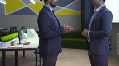 Ofiste takım elbiseli iki adam konuşuyor, müzakere ediyor, toplantı ve iş hakkında konuşuyor. Arka planda, modern bir ofis, masalar, bilgisayarlar..