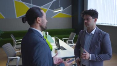 Two man employees in suits are talking in the office, a boss and a worker, solving work tasks. A man is holding a laptop.